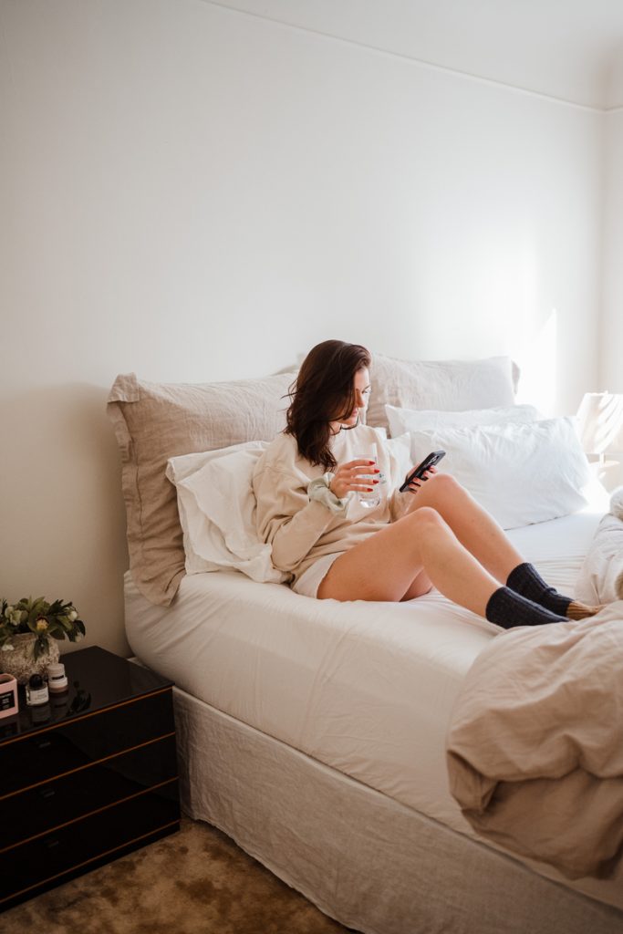 Carrie Barber sits in bed drinking a glass of water and reading on her phone_how to wake up easier