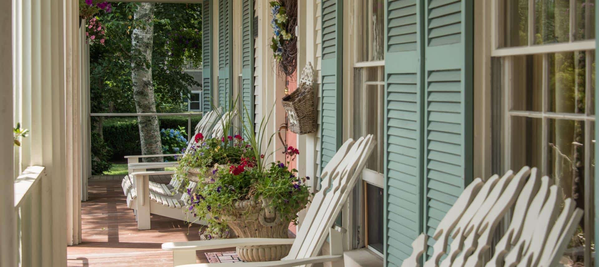 Front porch with multiple cream colored Adirondack chairs, potted planters with pink and purple flowers, salmon colored front door, and light green shutters