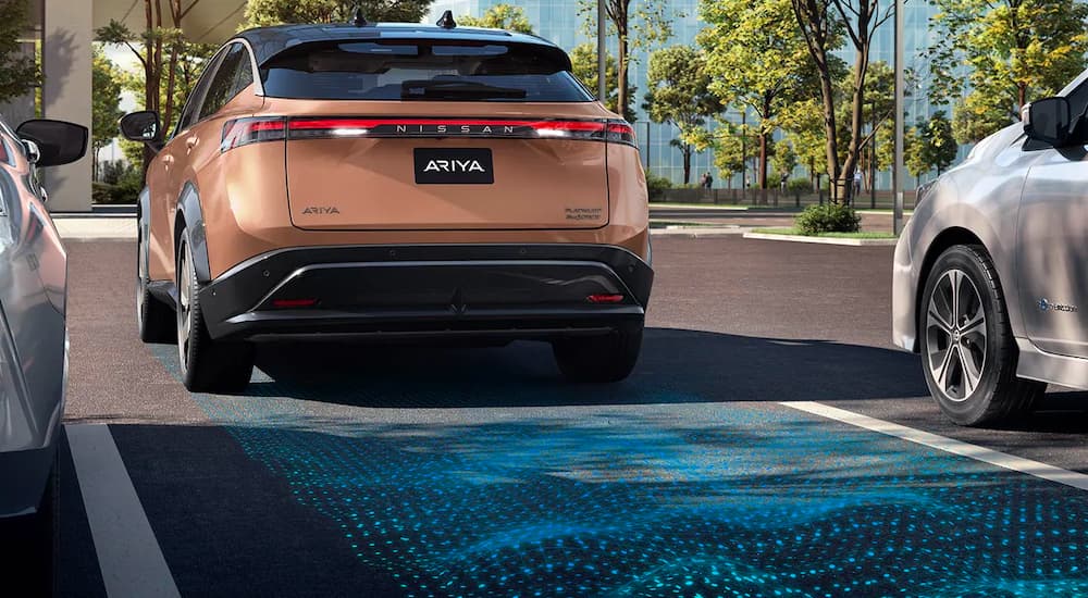 A bronze 2023 Nissan Ariya is shown from the rear while backing into a parking space after leaving a Nissan dealer.