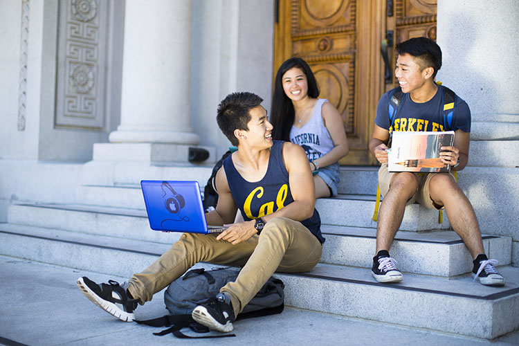  Three students sit on the steps of a building chatting, wearing Cal t-shirts, and holding their laptops.