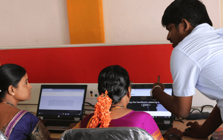 In a computer lab, an instructor speaks to three students who are facing their laptops.