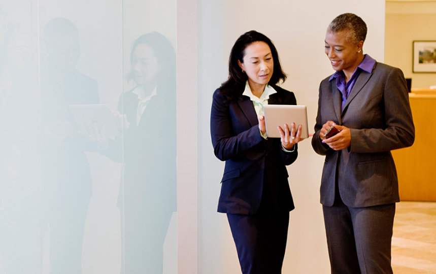 Two businesswomen dressed in suits are using a digital tablet while chatting in an office corridor. 