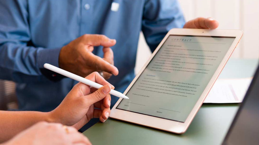 A seller holding a tablet with a short contract on display, while a customer is about to sign on it.