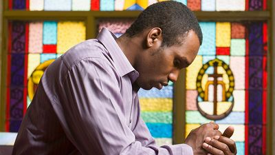 African American man praying in church with stained glass in background