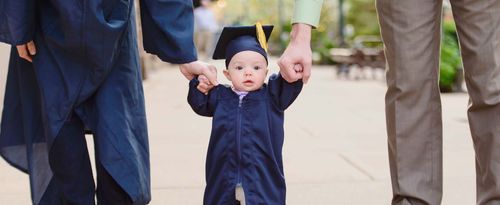 Baby preschool grad wearing cap and gown holding hands with mom and dad.