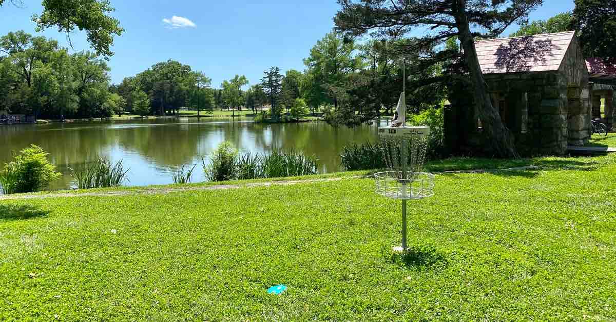 A disc golf basket in a green park area in fron of a pond