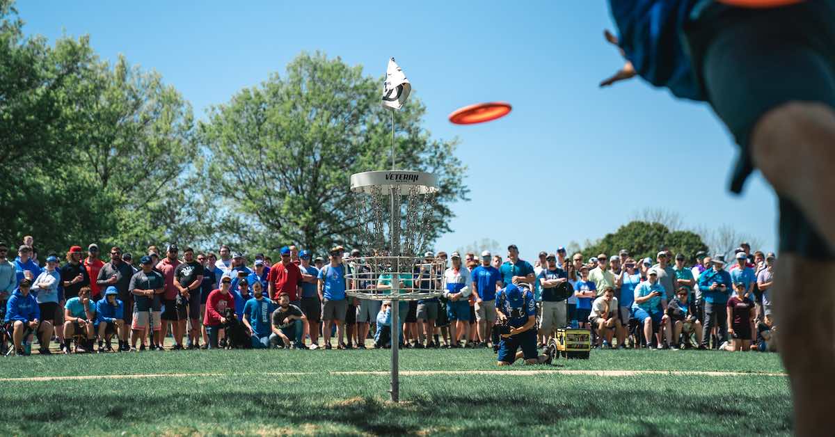 An orange disc golf putter sails toward a basket as a crowd watches on behind