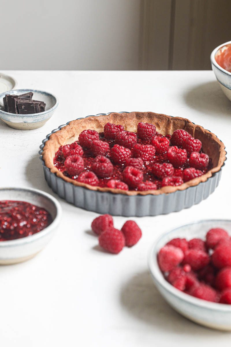 THe shortbread crust filled with the raspberry jam and fresh raspberries wtih a bwol of raspberrues on the front and a small bowl of chocolate in the back of the frame.