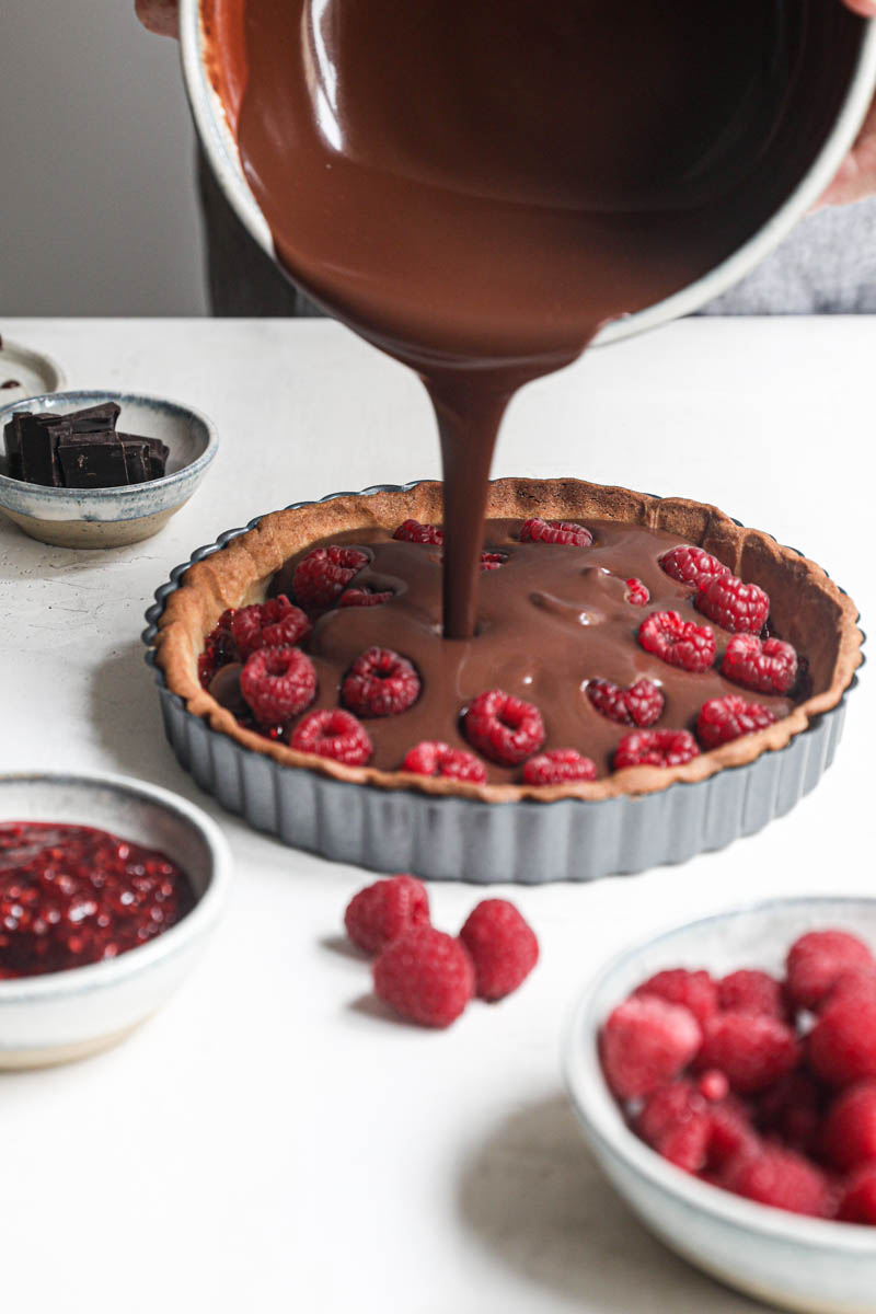 Two hands pouring the chocolate ganache over the shortbread crust filled with the raspberry jam and fresh raspberries wtih a bwol of raspberrues on the front and a small bowl of chocolate in the back of the frame.