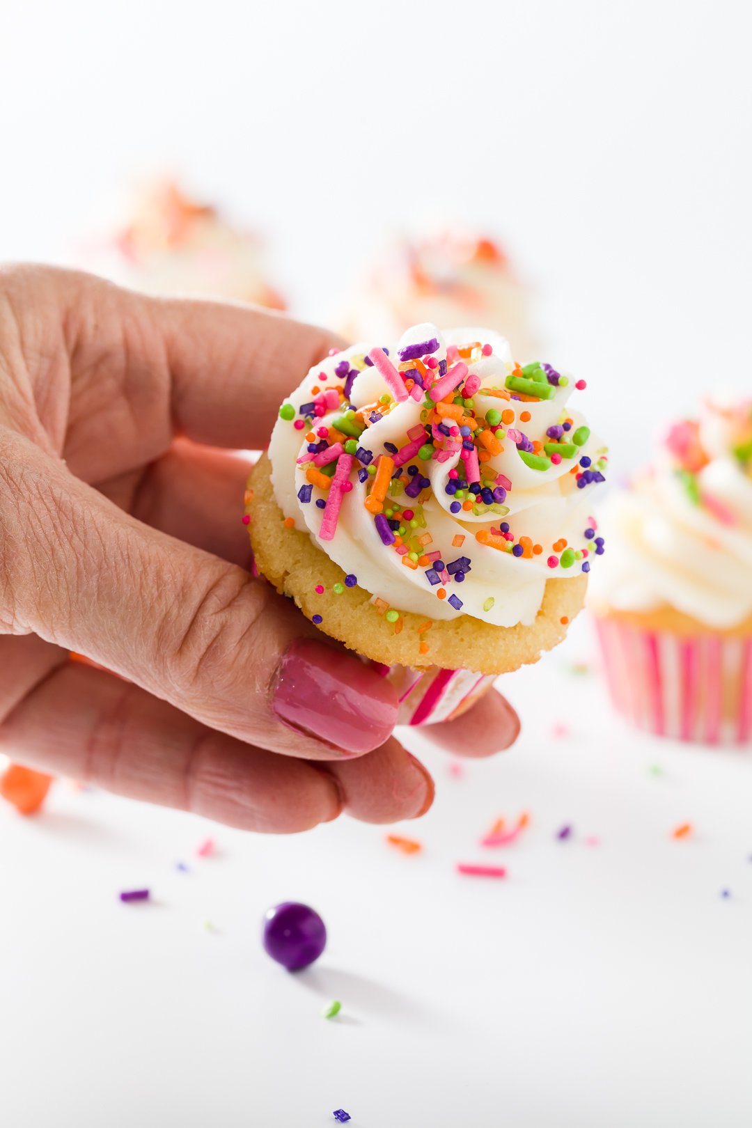 Stef's hand holding a mini vanilla cupcake frosted with vanilla buttercream and decorated with sprinkles