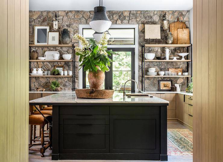 A stunning rustic stone wall accent an eclectic kitchen boasting wood and metal shelves fixed on either side of a black French doors located beneath a transom window. Burnt orange velvet stools sit at a dark brown wood island finished with a sink and lit by two Kelly Wearstler Precision pendants.