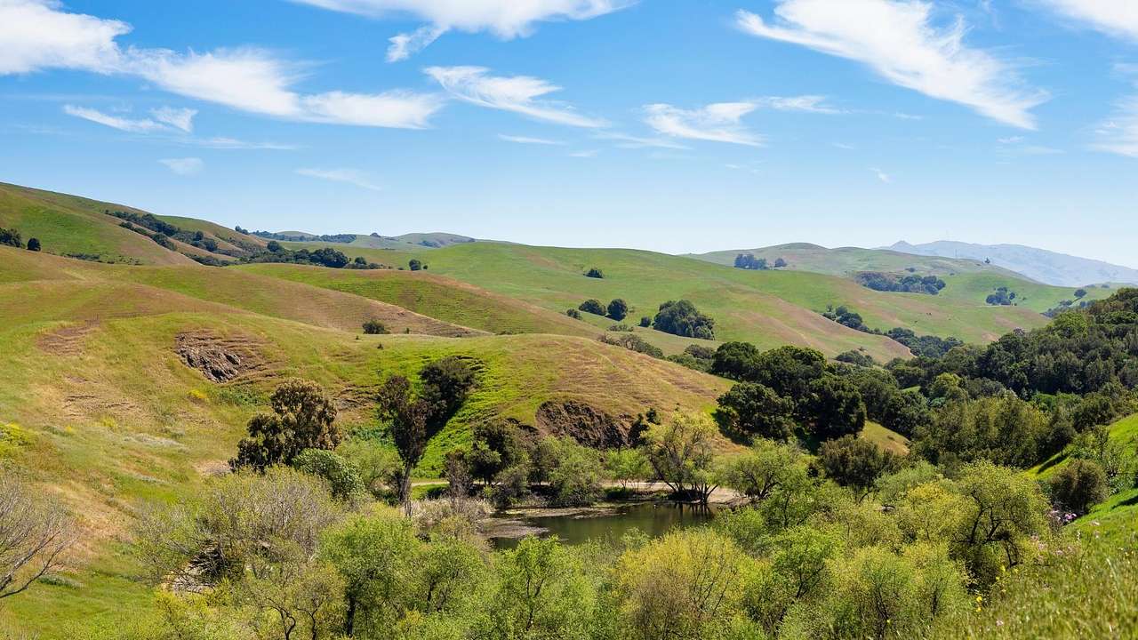 A view over greenery-covered hills and a small pond under a blue sky with clouds