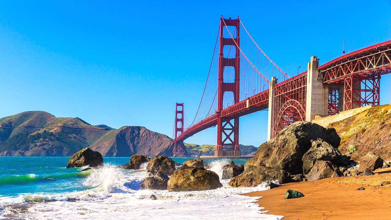 A red suspension bridge next to the ocean and a small beach with sand and rocks