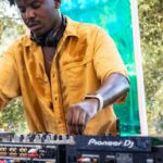 A young black DJ wearing headphones and a bright yellow button up shirt plays music on a Pioneer DJ pro set-up.