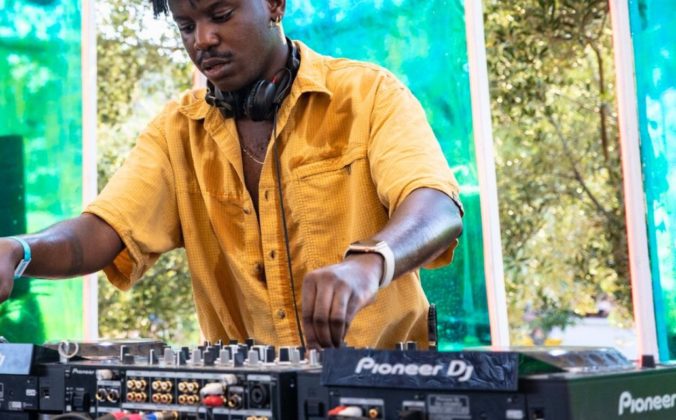 A young black DJ wearing headphones and a bright yellow button up shirt plays music on a Pioneer DJ pro set-up.