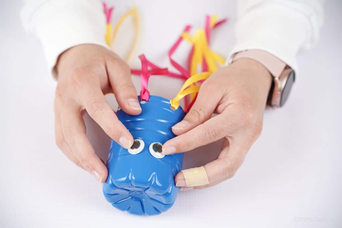 hand putting googly eyes onto top half of blue bottle windsock