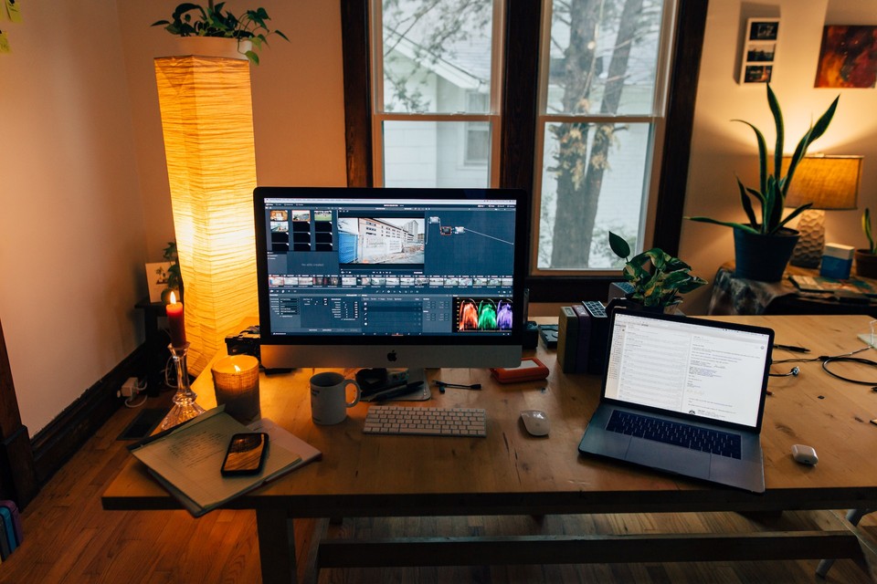 Home office setup with a desktop and laptop on a wooden table, surrounded by plants and a large window in the background