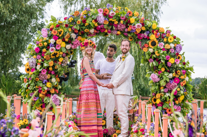 Bride wears bold pink wedding outfit and holds hands with groom at end of colorful and flowery aisle