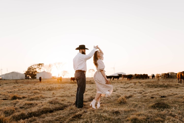 Western couple in cowboy hats twirl and dance on farm during sunset