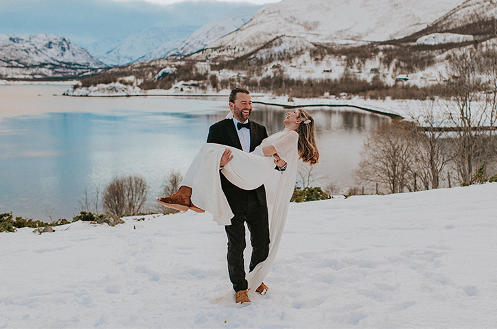 Man carrying woman celebrating their intimate elopement ceremony, in snowy landscape with lake and mountains in the background