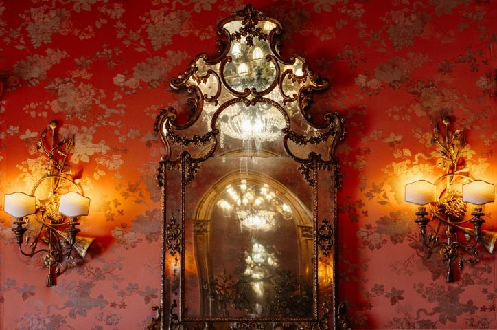 Vintage copper mirror with intricate detailing between wall sconces on patterned wallpaper wall