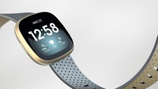 En Fitbit Versa 3 i ljusblått och med gulddetaljer mot en vit bakgrund.