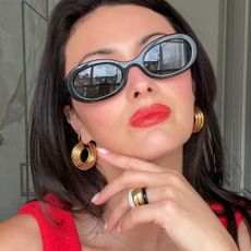 A close up of the face of a woman wearing black oval sunglasses with red lipstick and gold jewelery.