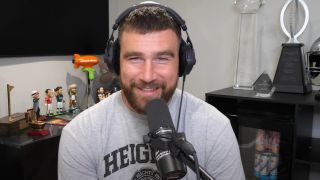 Travis Kelce smiling while wearing headphones and sitting behind a microphone during an episode of New Heights.