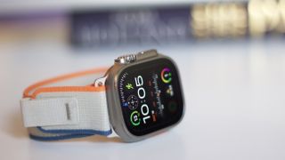 The Apple Watch Ultra 2 pictured on its side on a white table with a white and orange strap.