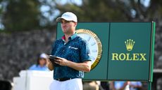 Justin Thomas looks at his yardage book with a large Rolex clock behind him at The Sentry 2025