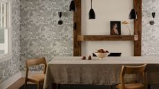 A dining table with fruits in a bowl. Wooden features and textures are seen throughout the room. Black pendant lighting hangs above the dining table as light floral wallpaper is wrapped around the rooms walls 