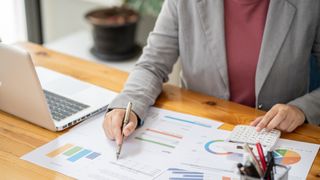 Businessperson using calculator and looking at financial charts with laptop by their side