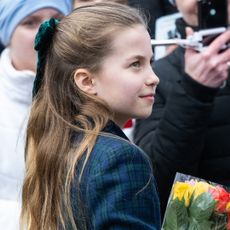 Princess Charlotte wearing a blue and green plaid coat and green hairbow holding flowers and smiling while looking to the right