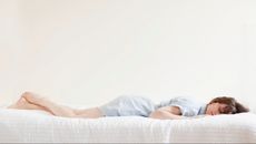 Person wearing light blue pyjamas laying on their front on beige bed showing body impressions on the mattress.