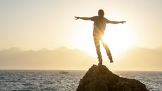 A person balances on one foot on a rock jutting out of the ocean, the sunset in the background.