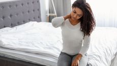 Woman sitting on the edge of her old mattress holding her neck in pain