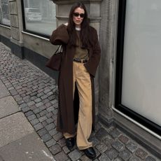 Annabel wears a brown long coat, brown sweater, black belt, tan trousers, and black boots.