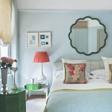 A bedroom painted in pale blue with a scalloped green mirror hanging above the bed