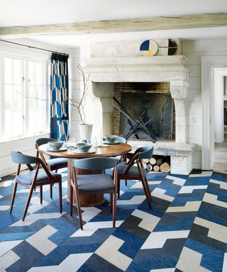 Traditional dining room space with white, panelled wooden walls, white painted ceiling, large stone fireplace, striking, geometric-patterned tile carpet flooring, blue, white and cream colorways, matching curtains, dark wood, rounded dining table, four dining chairs in dark wood with blue seat and back upholstery