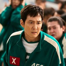 A man with a worried expression (Lee Jung-jae as Gi-hun) stands among a crowd of people in identical green tracksuits, in 'Squid Game' season 2.