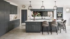 Classic style kitchen with white countertops, gray cabinets, pale gray tiles, bar stools, metal and glass pendant lights, mirror, vase of flowers,