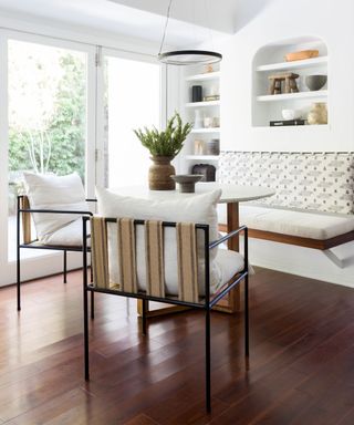 Relaxed dining room space with white painted walls, dark wood flooring, recessed, arch shelving spaces, decorated with unique ornaments and objects, wall mounted bench with upholstered backrest, two webbed dining chairs with black metal frames and cream cushions