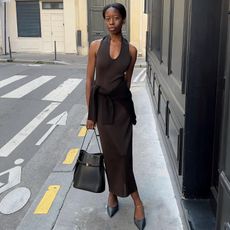 French woman wears a Khaite dress with a sweater tied around her waist, pointy toe heels and carrying a Toteme bag.