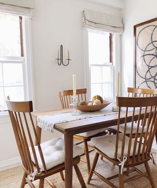 White dining room with wooden dining table and traditional, wooden dining chairs with seat cushions, cream table runner with candles and wooden fruit bowl, metal candelabra on wall, large, abstract artwork