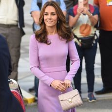 Kate Middleton walking outside with people behind her taking photos while wearing a purple long-sleeved dress holding a lilac top-handled Aspinal bag 