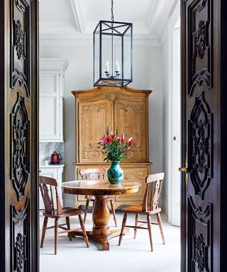 Looking through open ornate period mahogany doors into kitchen with country style round table and chairs and walnut dresser