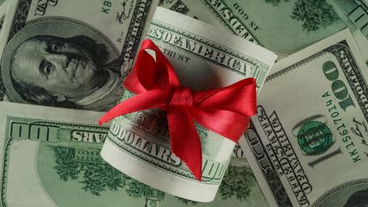 A roll of cash wrapped with a red ribbon against a background of hundred-dollar bills. 