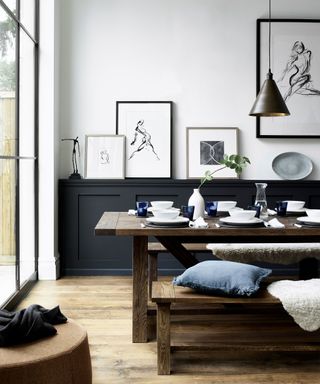 White dining room with black wall paneling half-way up the wall, large, dark wood dining table with benches with seat cushions, ornaments, frames and accessories placed on paneling shelf, dark wooden flooring, low hanging cone pendant above dining table