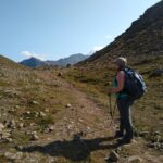 A woman stands on a trail looking at mountains in the distance. She has a blue backpack and is wearing a hat and sunglasses. She is using hiking poles and wearing gray long pants and a light green tank top