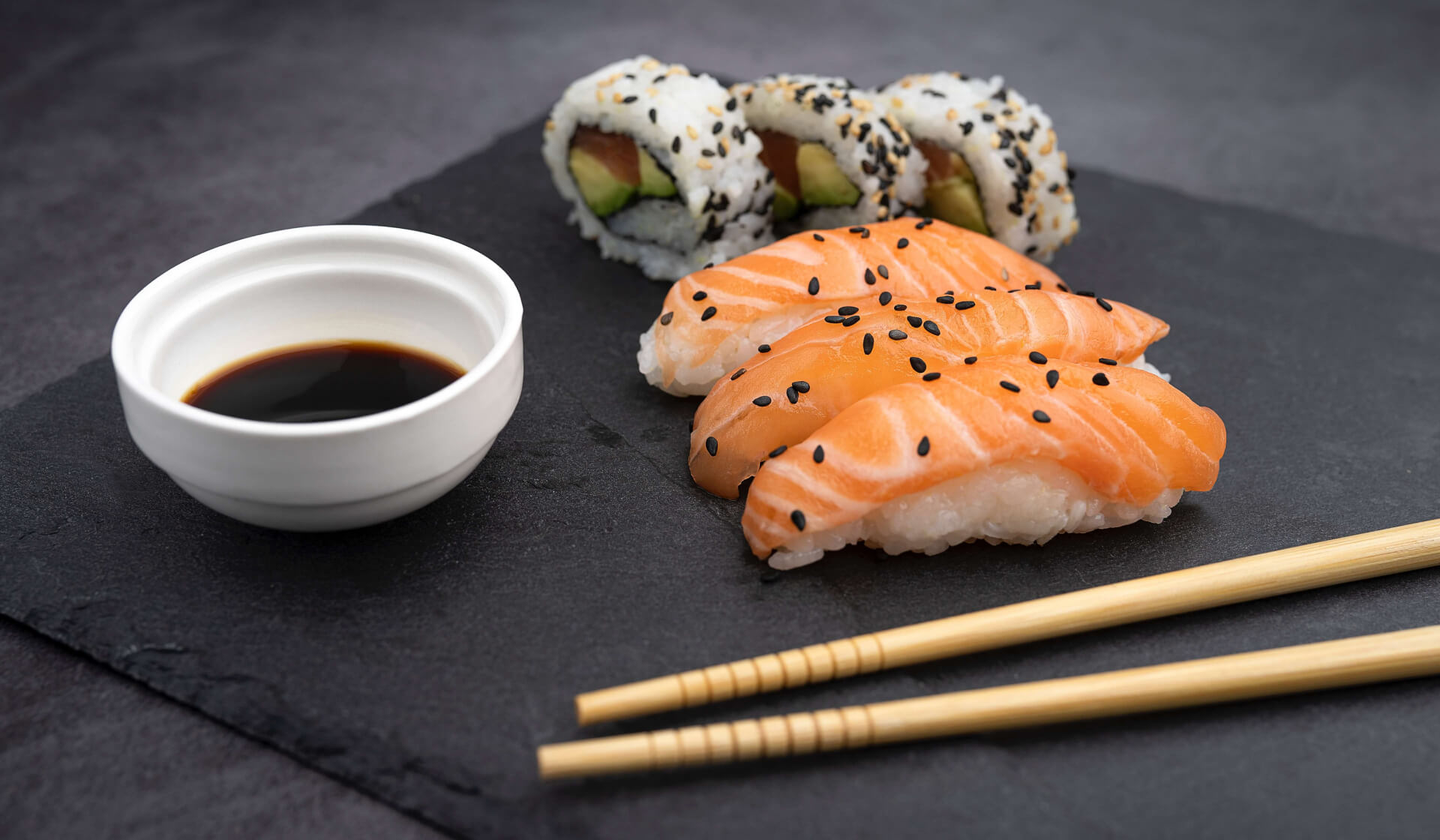 Japanese salmon sushi and a tuna avocado roll on a slate with soy sauce and chopsticks